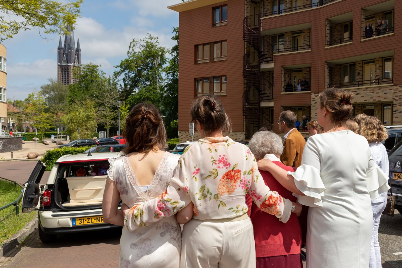 Familie staat arm in arm bij de Minirouwauto met open achterklep, buren kijken mee vanaf hun balkon